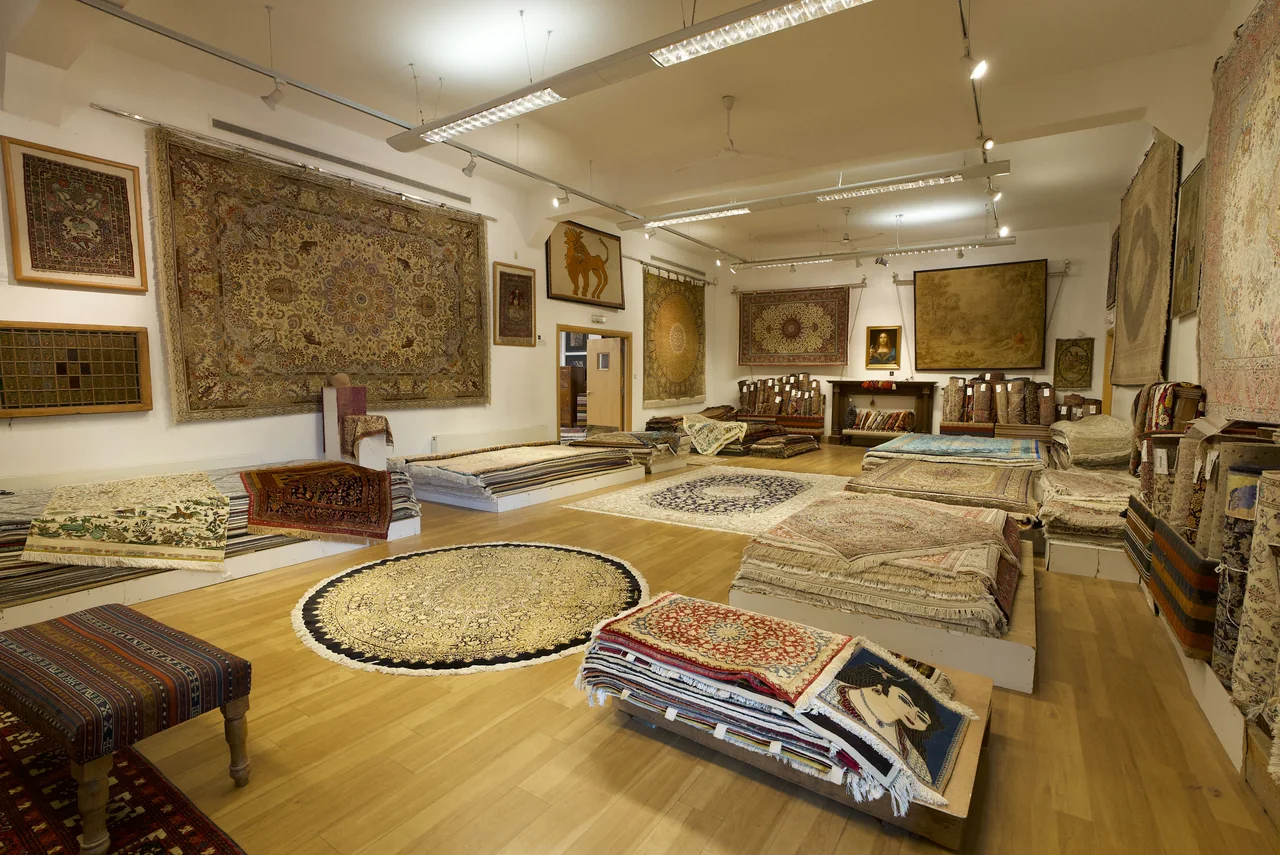 A smaller showroom room with a wooden floor, a round rug and fine carpets on the walls